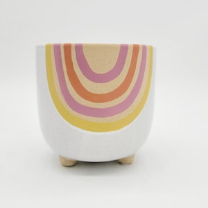 May include: White ceramic planter with a speckled finish, featuring a colorful rainbow design in yellow, pink, and orange. The planter has three small wooden feet. A decorative item for plants.