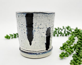 Puede incluir: Una taza de cerámica blanca con diseños abstractos negros y azules. La taza tiene una textura moteada.