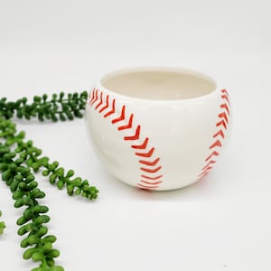 May include: White ceramic planter shaped like a baseball with red stitching. The planter is empty and sitting on a white surface.