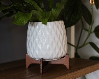 May include: A white ceramic planter with a diamond pattern sits on a wooden stand. The planter is filled with a green leafy plant. The planter is on a wooden shelf with a gold figurine and a white vase.