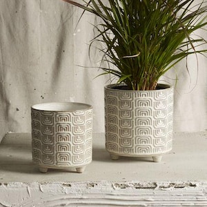 May include: Two white ceramic planters with a geometric pattern, one holding a tall green plant. The planters are on a white textured surface. The smaller planter is empty. The larger planter has a plant with green and brown leaves.