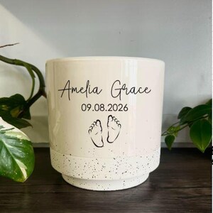 May include: A cylindrical ceramic planter with a cream-colored upper section and a white base featuring black speckles. The front displays the black text Amelia Grace, the date 09.08.2026, and a graphic illustration of two small baby footprints.