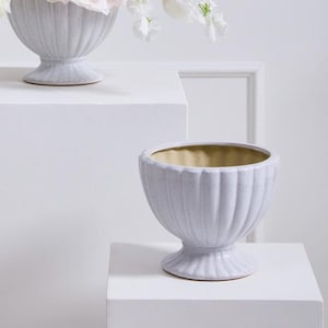 Puede incluir: Arreglo floral con flores blancas, como peonías y ranúnculos, en un jarrón gris claro. También hay un segundo jarrón similar. El arreglo se exhibe sobre un pedestal blanco contra un fondo blanco.