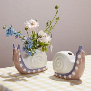 May include: Two snail-shaped ceramic vases. One vase holds a bouquet of flowers. The vases are white with brown and purple accents and white polka dots. The vases sit on a yellow and white checkered tablecloth.