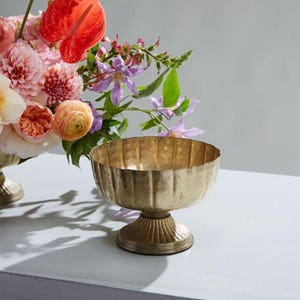 May include: A gold-colored, pedestal bowl with a textured surface sits next to a floral arrangement. The arrangement features pink, peach, and red flowers, including dahlias and anthuriums. The background is a light gray.
