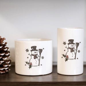 Könnte beinhalten: Zwei weiße Keramik-Kerzenhalter mit einem Schneemann-Design. Der Schneemann trägt einen Zylinder und einen Schal und hält einen Besen. Links befindet sich ein Tannenzapfen. Die Kerzenhalter sind zylindrisch und stehen auf einem dunkelbraunen Regal.