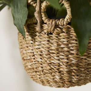 May include: Close-up of a woven basket with a round handle and a natural, light brown color. The basket is filled with green leaves. The basket is made of woven material and has a textured surface.