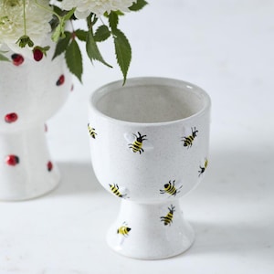 May include: White ceramic pedestal planter with a speckled finish, adorned with black and yellow bee illustrations. The planter is designed with a wide opening and a rounded base. The image also shows a second planter and white flowers.