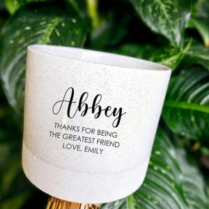 May include: A white cylindrical planter with a speckled finish featuring the black printed text Abbey, THANKS FOR BEING THE GREATEST FRIEND, LOVE, EMILY. The planter has a smooth rim and a slightly tapered base, presenting a clean and simple design.