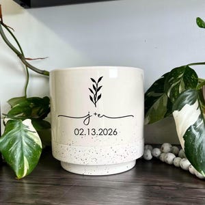 May include: A cylindrical ceramic planter with a cream-coloured finish and a speckled base. The front features a black graphic of a leafy sprig above the text j+e and the date 02.13.2026 in a clean, sans-serif font centred on the smooth surface.