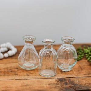 May include: Three clear glass bud vases of different shapes are arranged on a wooden surface. The empty vases have a vintage appearance. The background is a neutral grey, suggesting a focus on home decor or floral arrangements. The vases are empty.
