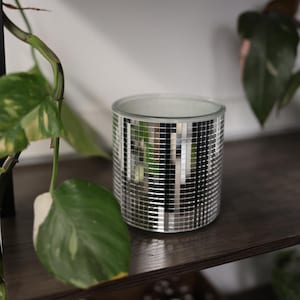 May include: A cylindrical candle holder with a mirrored mosaic tile design. The silver tiles reflect light, creating a shimmering effect. The candle holder sits on a dark wooden shelf, with green plants in the background.