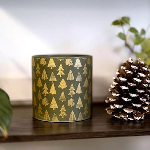 Könnte beinhalten: Zylindrischer, olivgrüner Keramik-Pflanztopf mit einem sich wiederholenden goldenen Baummuster. Rechts befindet sich ein großer Tannenzapfen mit weißen Akzenten. Der Topf steht auf einem dunkelbraunen Holzregal. Der Topf hat einen Durchmesser von ca. 10 cm.