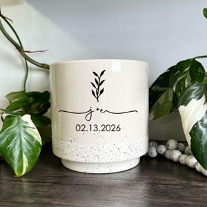 Op de afbeelding: Een cilindervormige keramische plantenpot met een crèmekleurig bovendeel en een gespikkelde wit-grijze basis. De voorzijde toont een zwarte afbeelding van een takje boven de tekst j+e en de datum 13.02.2026 in een eenvoudig zwart lettertype.