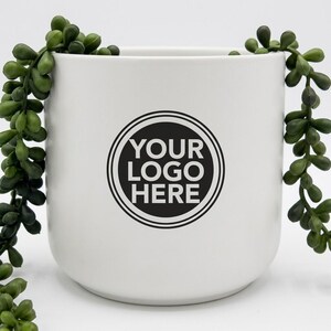 Pode incluir: Um vaso cilíndrico de cerâmica branca com um emblema circular preto na frente contendo o texto YOUR LOGO HERE em branco. O vaso é decorado com trepadeiras suculentas verdes que pendem de ambos os lados do recipiente.