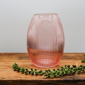 Może przedstawiać: Różowy szklany wazon z pionowymi żebrowanymi detalami. Wazon ma zaokrąglony kształt i stoi na drewnianej powierzchni. Zielone, liściaste elementy dekoracyjne są ułożone wokół wazonu.