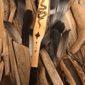 Op de afbeelding: Een houten toverstaf met een zwarte handvat en een slangontwerp gebrand in het hout. De toverstaf heeft een zwarte lederen wikkel om het handvat en een kleine ruitvorm met een stip in het midden gebrand in het hout. De toverstaf is omgeven door veren.