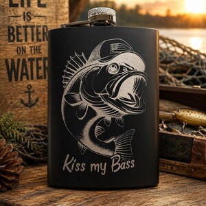 Op de afbeelding: Een matzwarte metalen heupfles met een lasergravure van een baars die een honkbalpet met een Amerikaanse vlag draagt. Onder de vis staat de tekst "Kiss my Bass" in een strak, wit en gestileerd lettertype geschreven.
