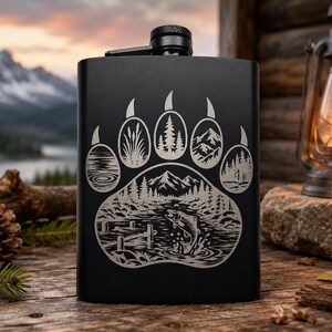 May include: A matte black metal flask featuring a laser-engraved design of a large bear paw print. Inside the paw print are detailed illustrations of a mountain landscape, a forest, a river, and a fish jumping near a wooden dock.