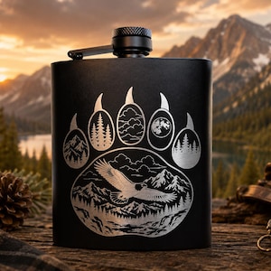 May include: A matte black metal flask featuring a laser-engraved bear paw print design. The paw pads contain illustrations of pine trees, clouds, and a full moon, while the palm area depicts an eagle flying over a mountain range and forest.