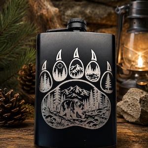 May include: A matte black metal flask featuring a laser-engraved design of a large bear paw print. Inside the paw print, there is a detailed landscape illustration of a bear walking through a forest with mountains in the background.