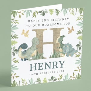 May include: A white card with a green leafy border. The card says "Happy 2nd Birthday to our roarsome son" with a large brown letter H and the name "HENRY" below. The date "14th February 2023" is at the bottom of the card. There are three dinosaurs in the image, a green T-Rex, a green Triceratops, and a yellow Pterodactyl.