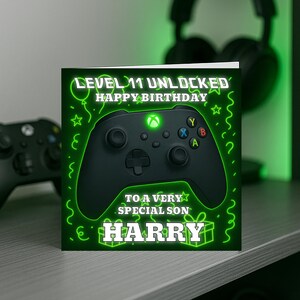 May include: A birthday card featuring a black Xbox controller design. The card's text reads "LEVEL 11 UNLOCKED HAPPY BIRTHDAY TO A VERY SPECIAL SON HARRY" in neon green. The background is green with neon green decorative elements.