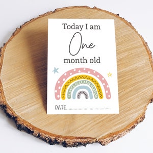 May include: A white card with the text "Today I am One month old" and a rainbow with yellow, pink, and blue colours. There is a space to write the date.