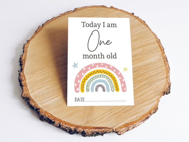 May include: A white card with the text "Today I am One month old" and a rainbow with yellow, pink, and blue colours. There is a space to write the date.