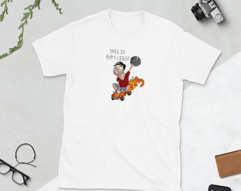 May include: White t-shirt featuring a cartoon illustration of a man riding a skateboard while holding a bowling ball. The text "THIS IS ROKT LEEG!!" is above the illustration. The man is wearing a red shirt and blue shorts.
