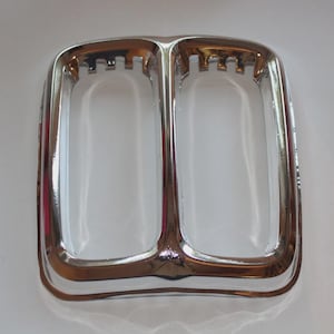 May include: A chrome ashtray with two compartments. The rectangular ashtray has a curved, Art Deco design and a shiny, reflective surface. The interior is clear, and the overall shape is symmetrical. The ashtray is likely made of metal.