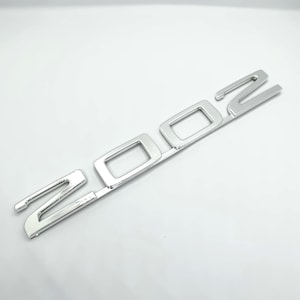 Peut inclure: Un emblème chromé brillant avec le chiffre "200". Les chiffres sont reliés par une fine barre. L'emblème a une surface réfléchissante, suggérant qu'il est en métal. Le design est moderne et épuré.