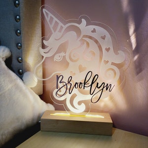 May include: A clear acrylic unicorn-shaped night light with the name "Brooklyn" in cursive script. The light sits on a wooden base, emitting a warm glow. The unicorn design includes hearts and swirls.