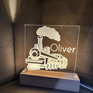 May include: A personalized night light featuring a train design with the name "Oliver" etched into a clear acrylic panel. The train illustration includes a smokestack and wheels, illuminated by a warm glow from the wooden base.