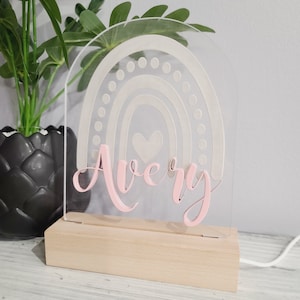 May include: A clear acrylic night light with a rainbow design and the name "Avery" in pink script. The light sits on a natural wood base. The rainbow features a heart in the center.