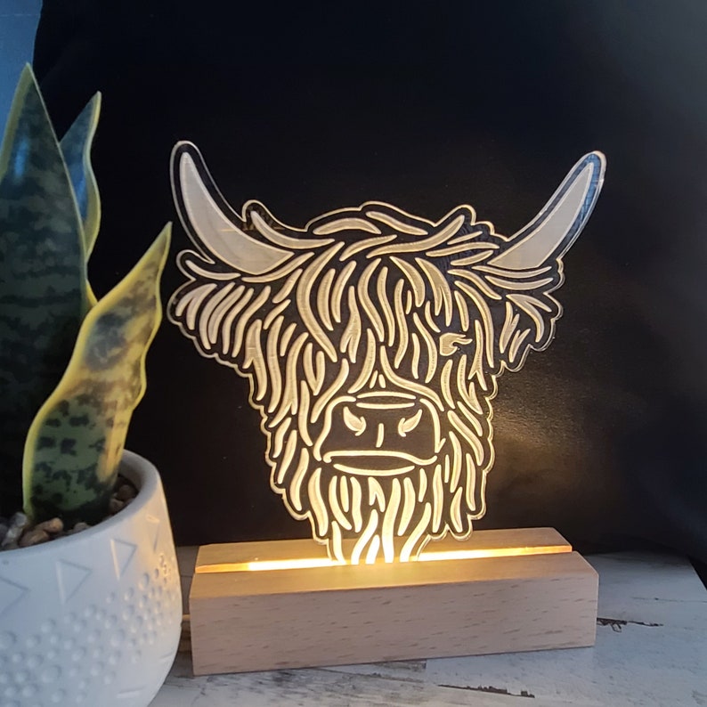 May include: A decorative Highland cow head-shaped night light. The light is made of clear acrylic with etched details, mounted on a wooden base. The light emits a warm glow, illuminating the cow's shaggy fur and horns. A potted plant is in the background.