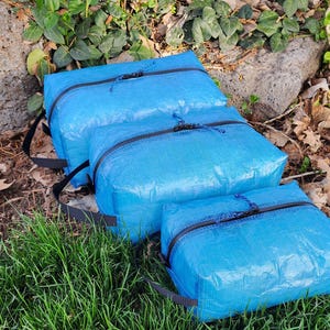 May include: Three rectangular storage pouches in bright blue, constructed from a textured, grid-patterned material. Each pouch features a black zipper closure with a blue cord pull and a black webbing handle attached to the side for carrying.