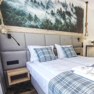 May include: A white and blue checked bedspread with white towels on a double bed in a hotel room. The headboard is light grey with a forest scene mural above it. There is a small wooden bedside table next to the bed.