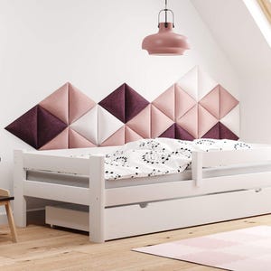 May include: White wooden bed with a pull-out drawer, a patterned comforter, and a geometric headboard in shades of pink and white. A small table and chair set are in the foreground, and a pink rug is on the floor.