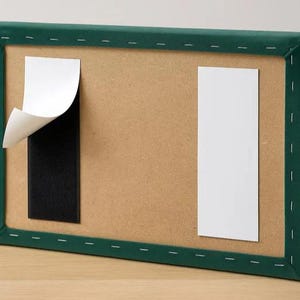 Puede incluir: Un lienzo rectangular con marco verde y un interior marrón. Dos tiras adhesivas rectangulares blancas están adheridas a la superficie marrón. Una tira tiene su respaldo parcialmente despegado, revelando una tira adhesiva negra.