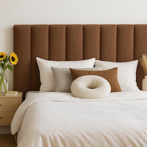 Op de afbeelding: Een slaapkamer met een bruin, verticaal bekleed hoofdbord. Het bed is opgemaakt met wit beddengoed en een selectie decoratieve kussens in verschillende kleuren en texturen. Links staat een klein houten nachtkastje met zonnebloemen in een vaas.