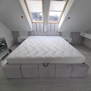 May include: A white upholstered bed frame with a white mattress and a quilted top. The bed frame has a storage compartment with a black pull strap.