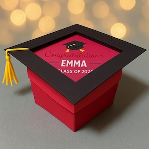 May include: A red graduation cap-shaped gift box with a black frame and a yellow tassel. The top of the box features the text "Congratulations EMMA CLASS OF 2024" and a graduation cap illustration. The box is on a gray surface.