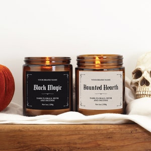 May include: Two amber glass candle jars with black labels. The labels have a decorative border and text that reads "Black Magic" and "Haunted Hearth". The candles are on a wooden surface with a white cloth and a small orange pumpkin.