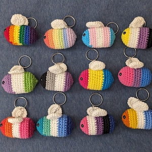 May include: Ten crocheted bee keychains with different coloured stripes. Each keychain has a white wing and a black eye. The keychains are hanging from silver key rings.