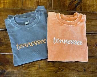 May include: Two long-sleeved shirts with the word "Tennessee" embroidered on the front. One shirt is grey and the other is orange.