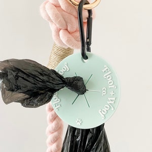 May include: A mint green dog waste bag dispenser with the text "Floof + Woof co." and a paw print. A black carabiner clip attaches the dispenser to a pink rope with a gold ring.
