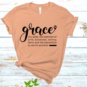 May include: A peach-colored t-shirt with the word "grace" in black cursive lettering. The word is defined as "the exercise of love, kindness, mercy, favor and the disposition to serve another".