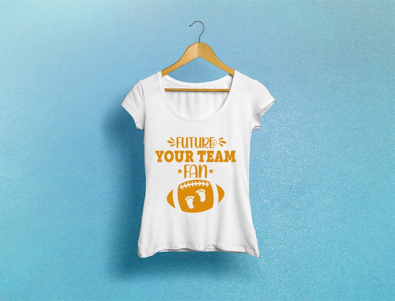 Puede incluir: Camiseta blanca con un gr&aacute;fico de f&uacute;tbol americano marr&oacute;n y el texto "FUTURE YOUR TEAM FAN" con una estrella a cada lado de la palabra "FAN".