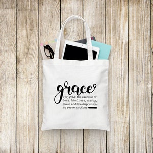 May include: White tote bag with black handles and the word "grace" written in cursive with a heart above the e. The definition of grace is written below in black text: "(m) gr&aring;s: the exercise of love, kindness, mercy, favor and the disposition to serve another."